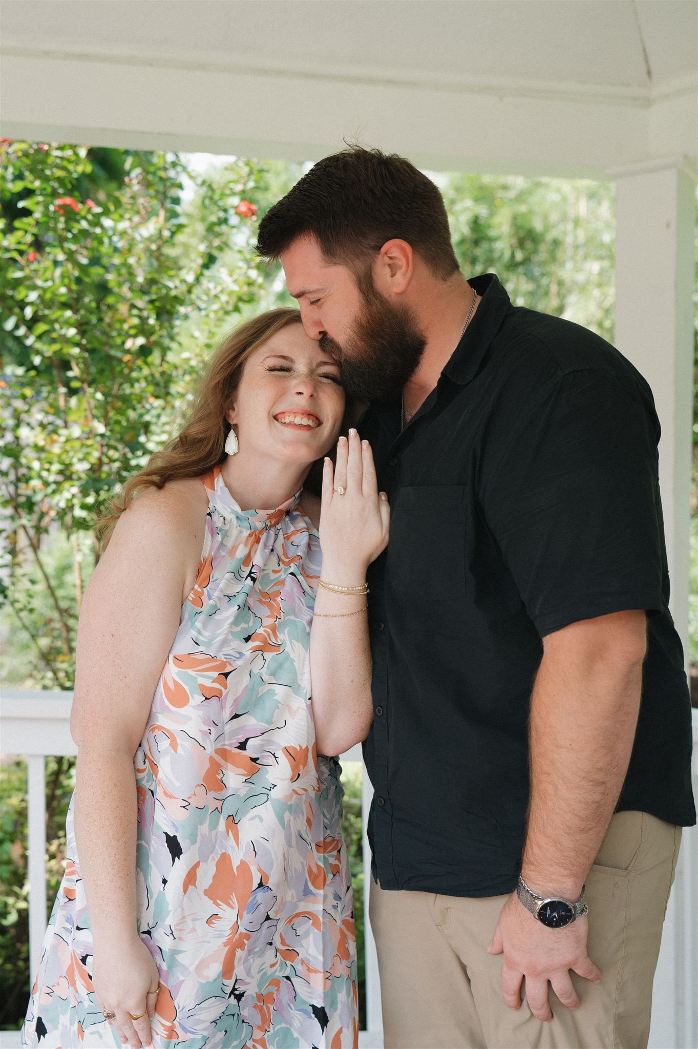 A couple shares an intimate moment on a white porch, with the woman wearing a floral dress and the man in a black polo.