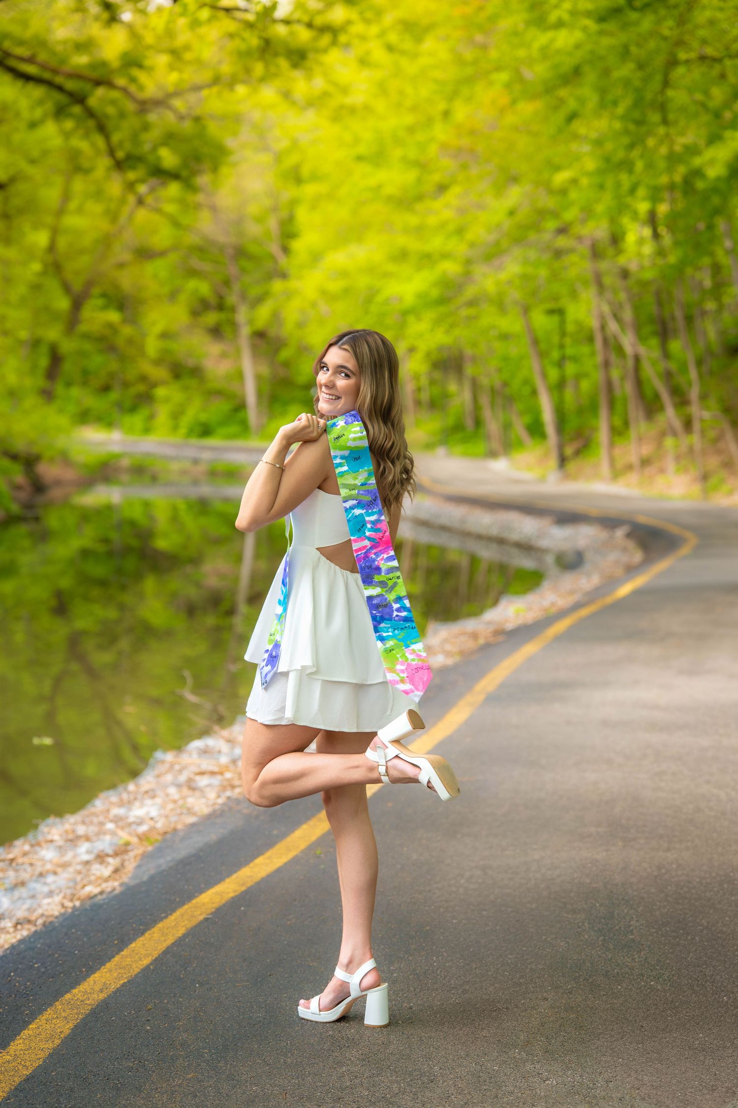 Young woman in white dress with colorful scarf posing on a scenic road surrounded by lush green trees near a pond.
