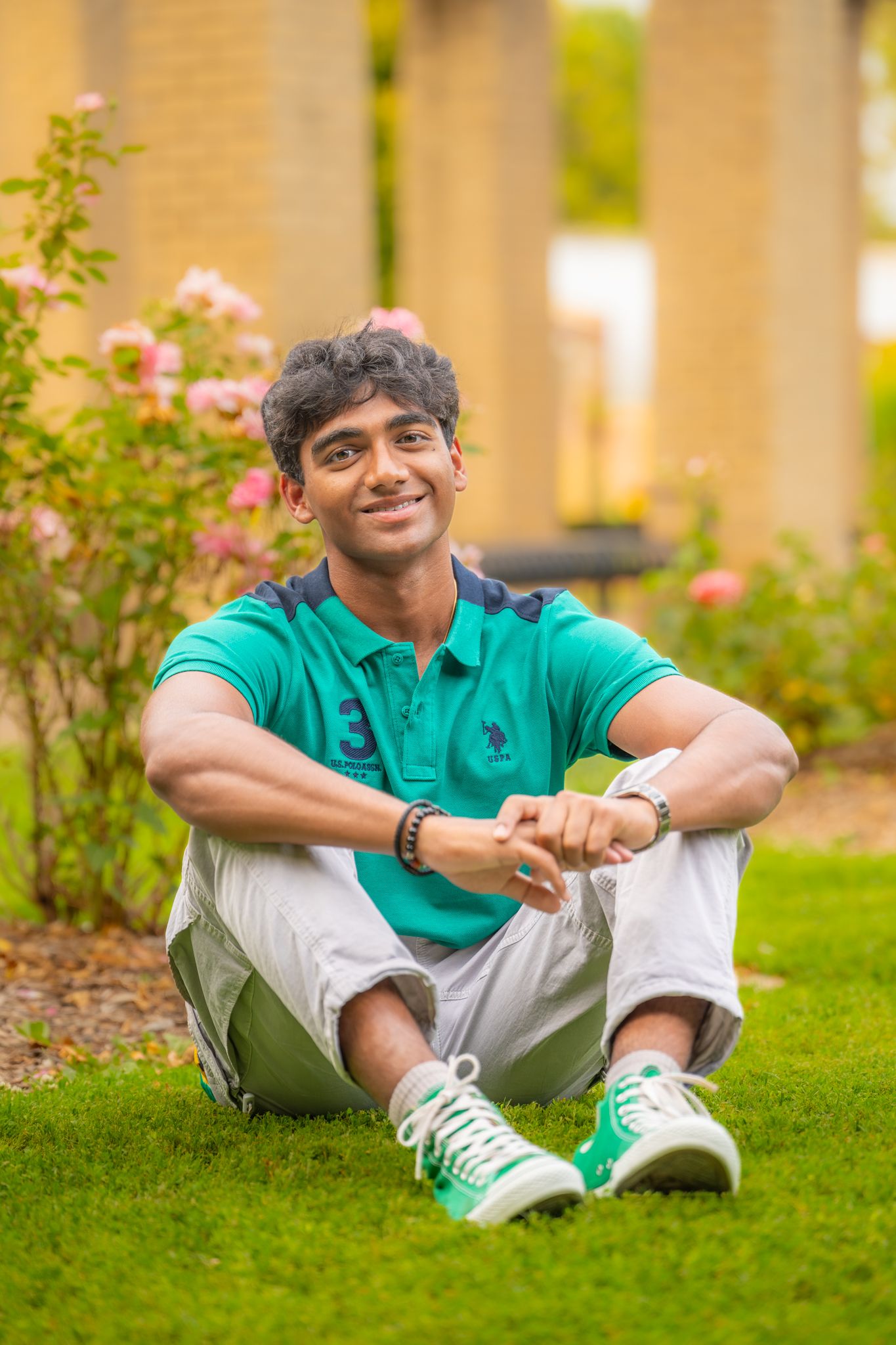 A person in a turquoise polo shirt and white pants relaxes on green grass in campus-style surroundings.