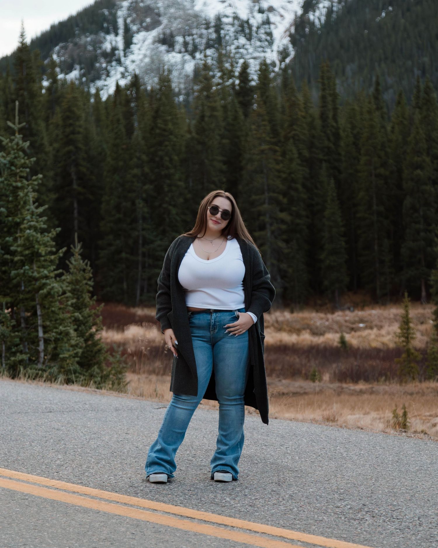 A person in casual wear poses confidently on a mountain road with evergreen forests in the background.