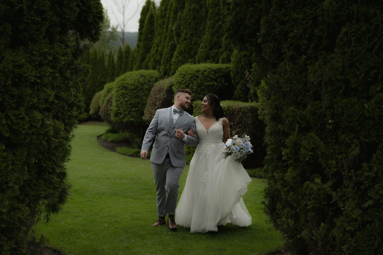 A couple walks together along a manicured garden path surrounded by evergreen trees.