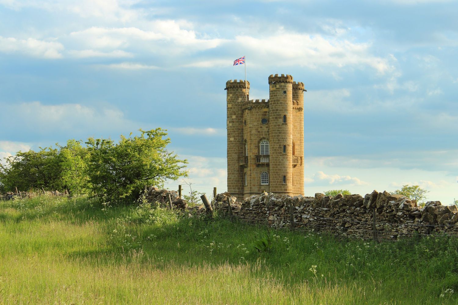 A scenic view of Broadway Tower, a historic folly atop the Cotswolds hills, surrounded by rolling countryside under a partly cloudy sky.