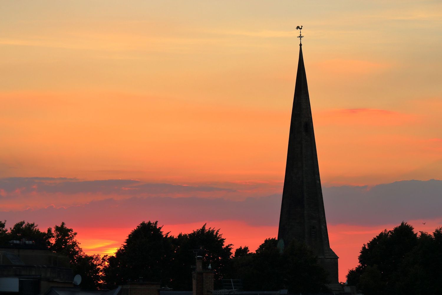 A majestic church steeple rises above the tree line, silhouetted against a stunning sunset. The warm hues of a orange, pink, and purple sky