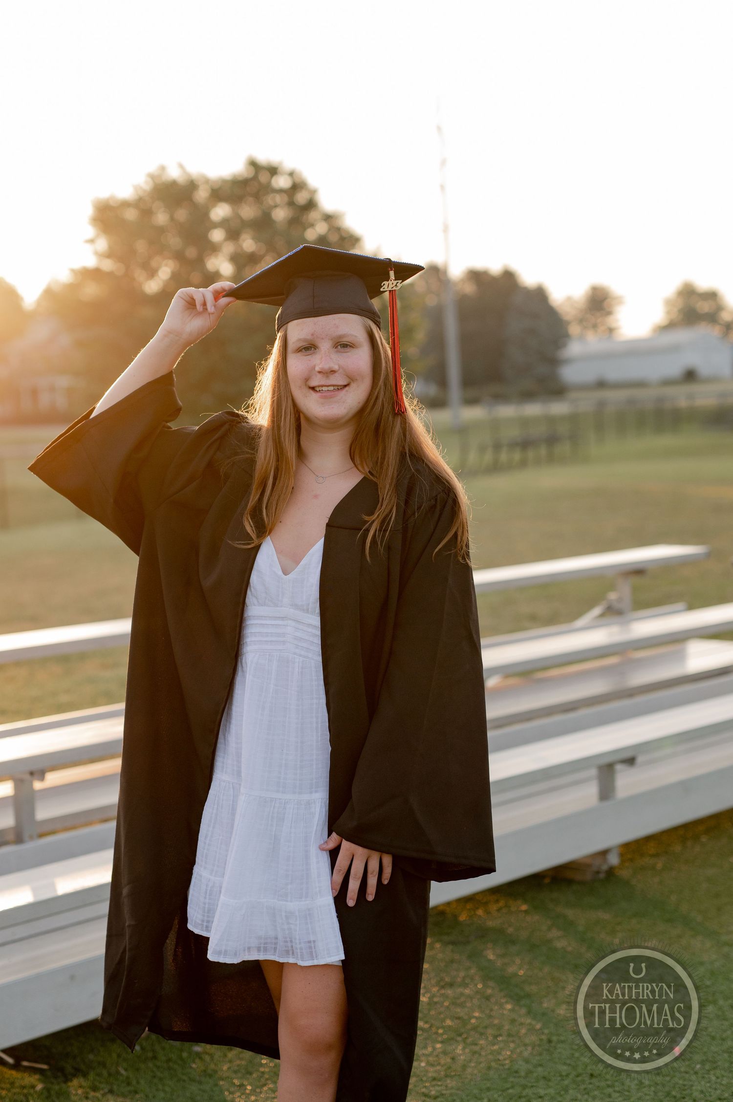Graduate in black cap and gown smiling on bleachers during sunset.