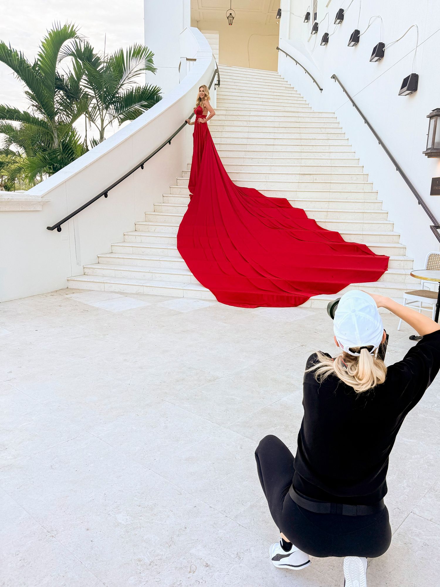 A photographer captures a dramatic red flowing dress on a grand white staircase with palm trees in the background.