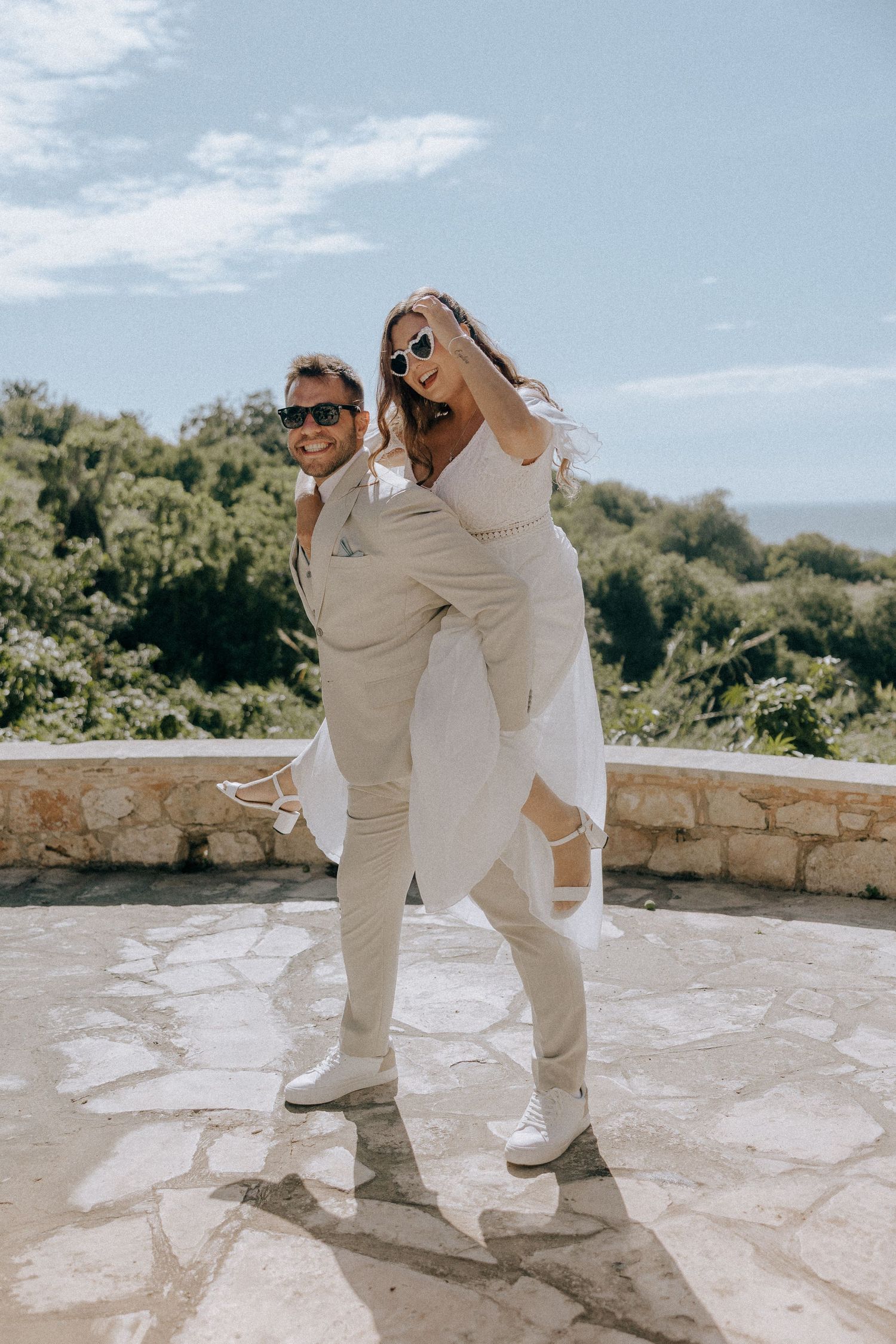 A joyful couple in matching beige outfits pose playfully outdoors against a scenic mountain backdrop.