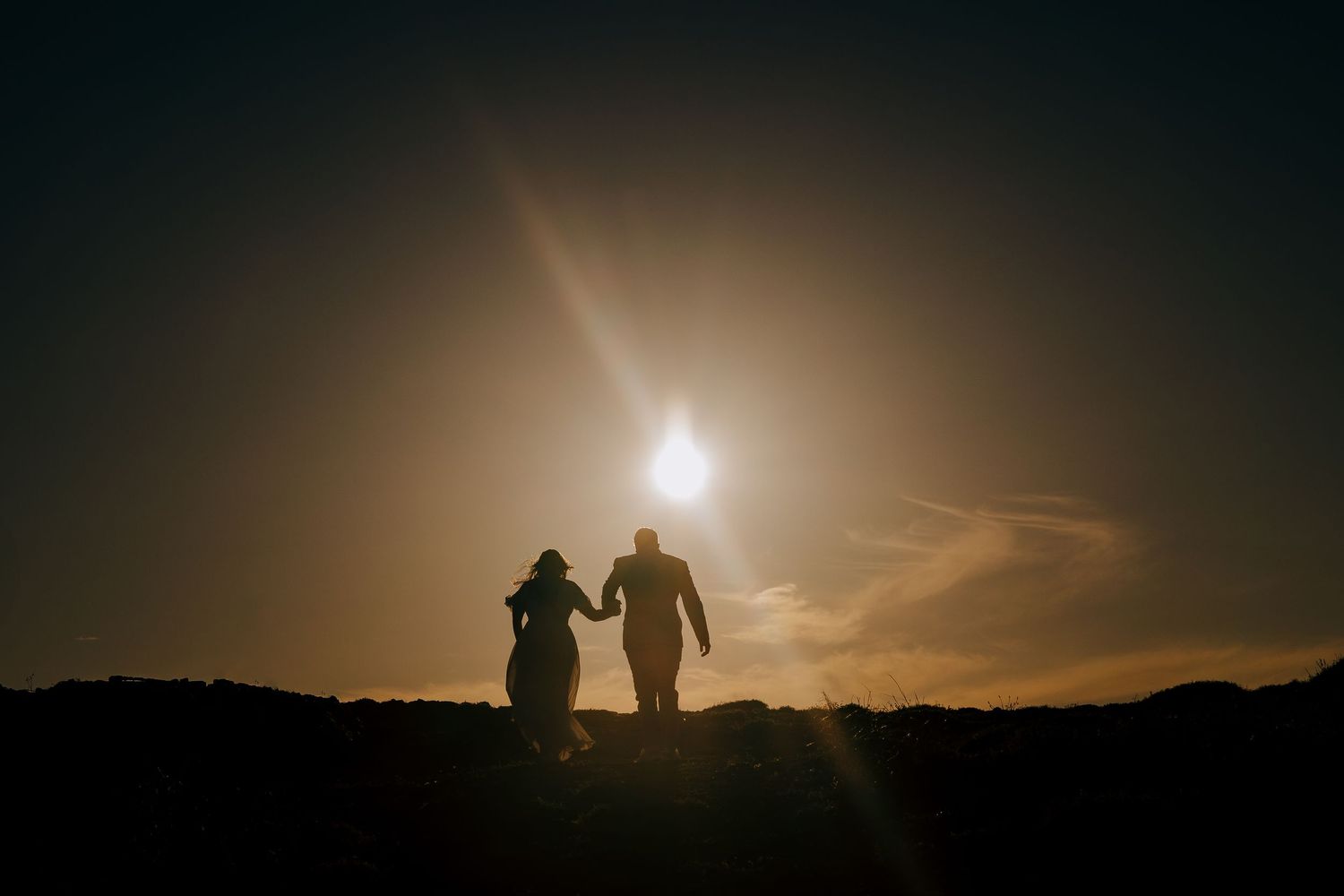 Two silhouetted figures walk hand in hand on a hilltop against a dramatic golden sunset sky.