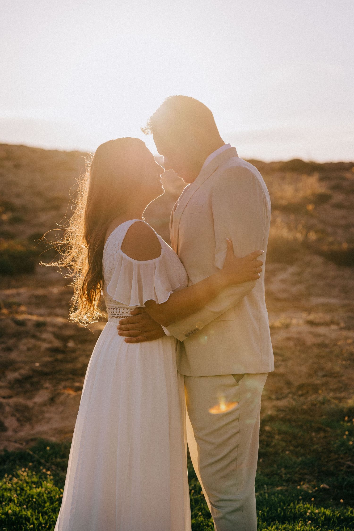 A couple in white embraces during a romantic sunset photoshoot in a natural outdoor setting.