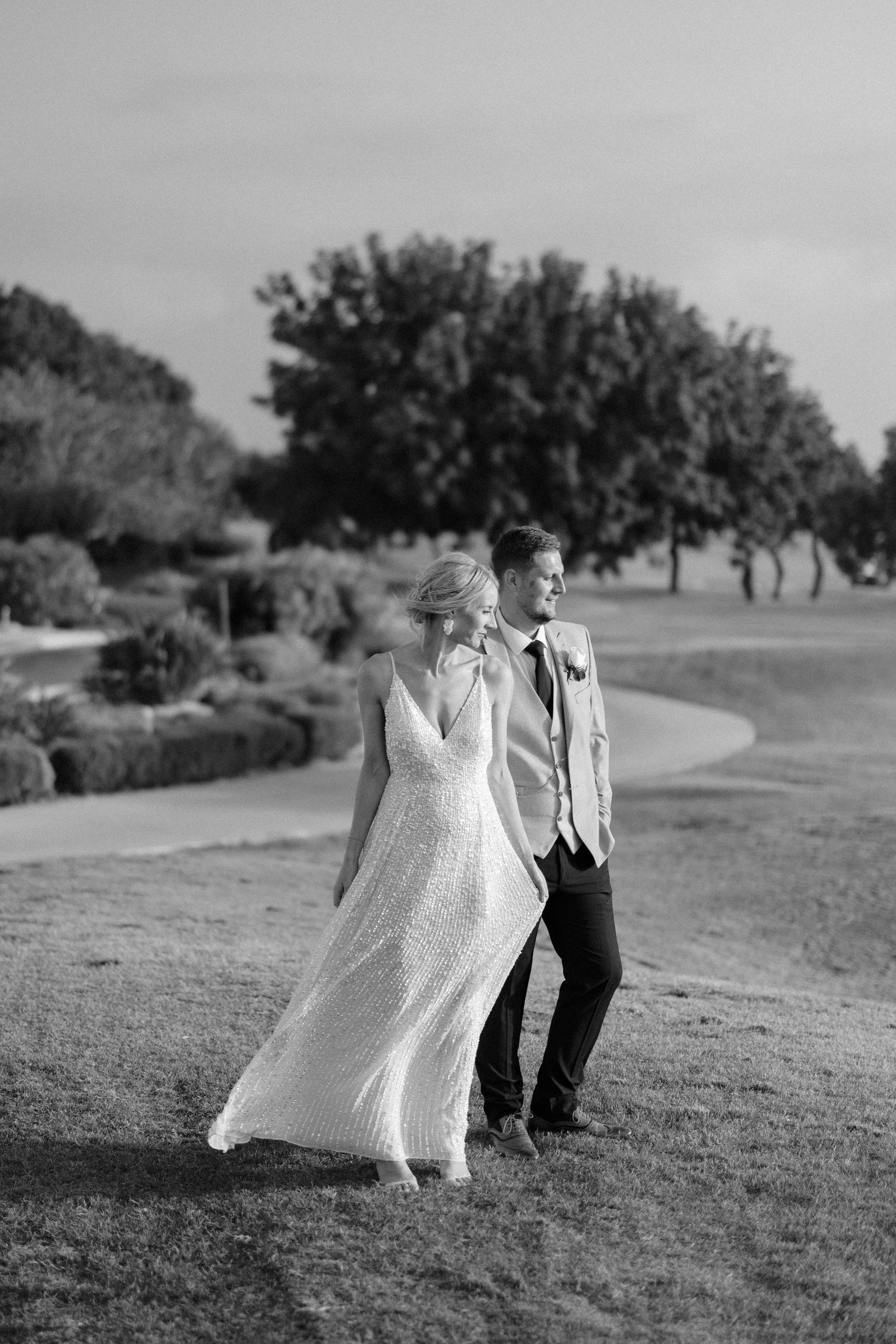 Black and white photo of a bride and groom walking outdoors on a golf course, bride in flowing gown, groom in suit.