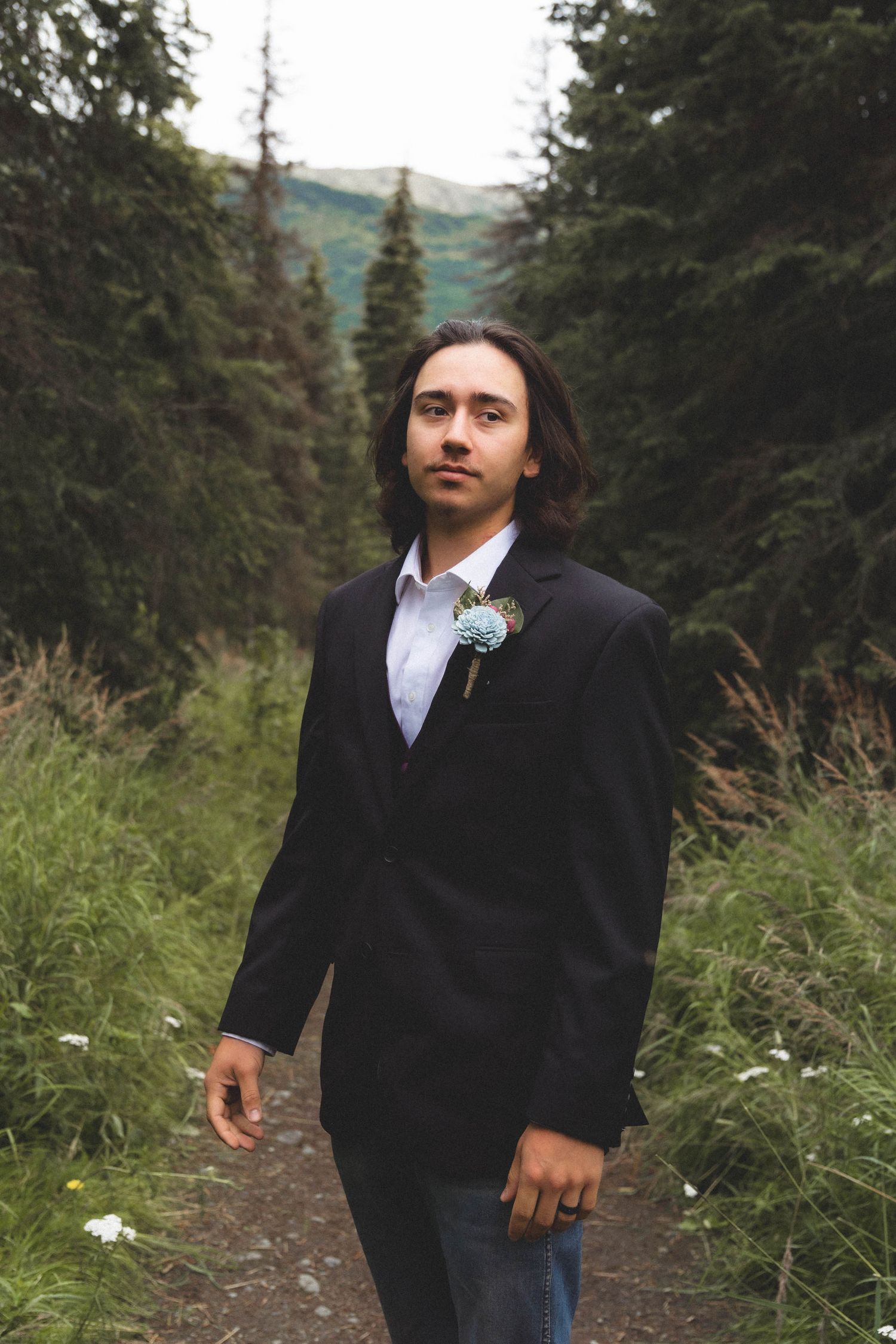 A groom in a black suit with a boutonniere stands on a forest path surrounded by pine trees.