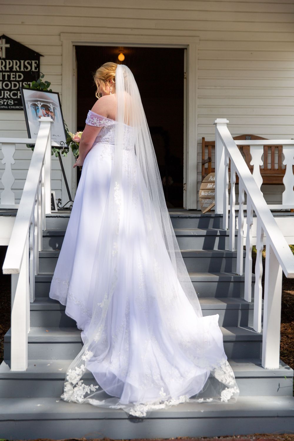 Bride in flowing white gown and veil standing on steps of white chapel building.