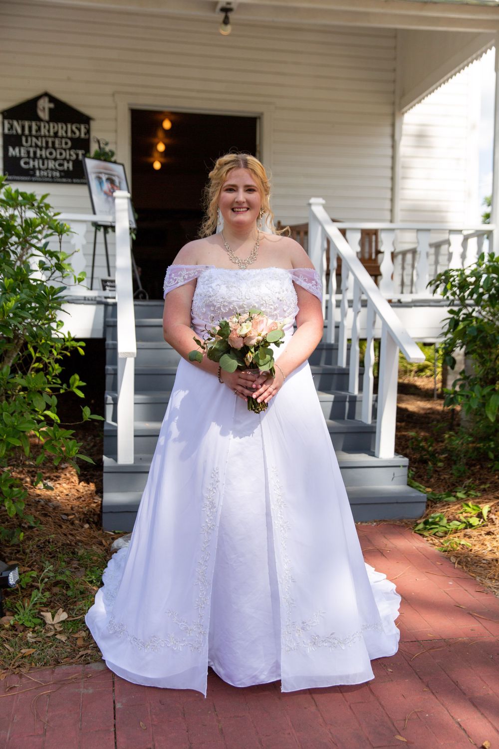 Bride in elegant off-shoulder gown posing with bouquet on church entrance steps.