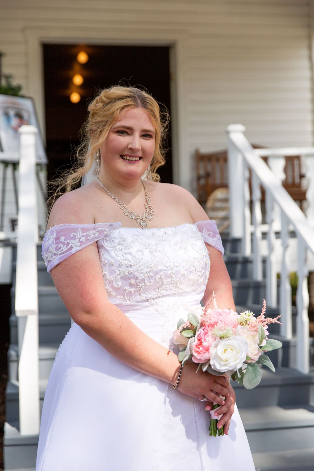 Bride wearing off-shoulder wedding dress holding pink and white bouquet on porch steps.