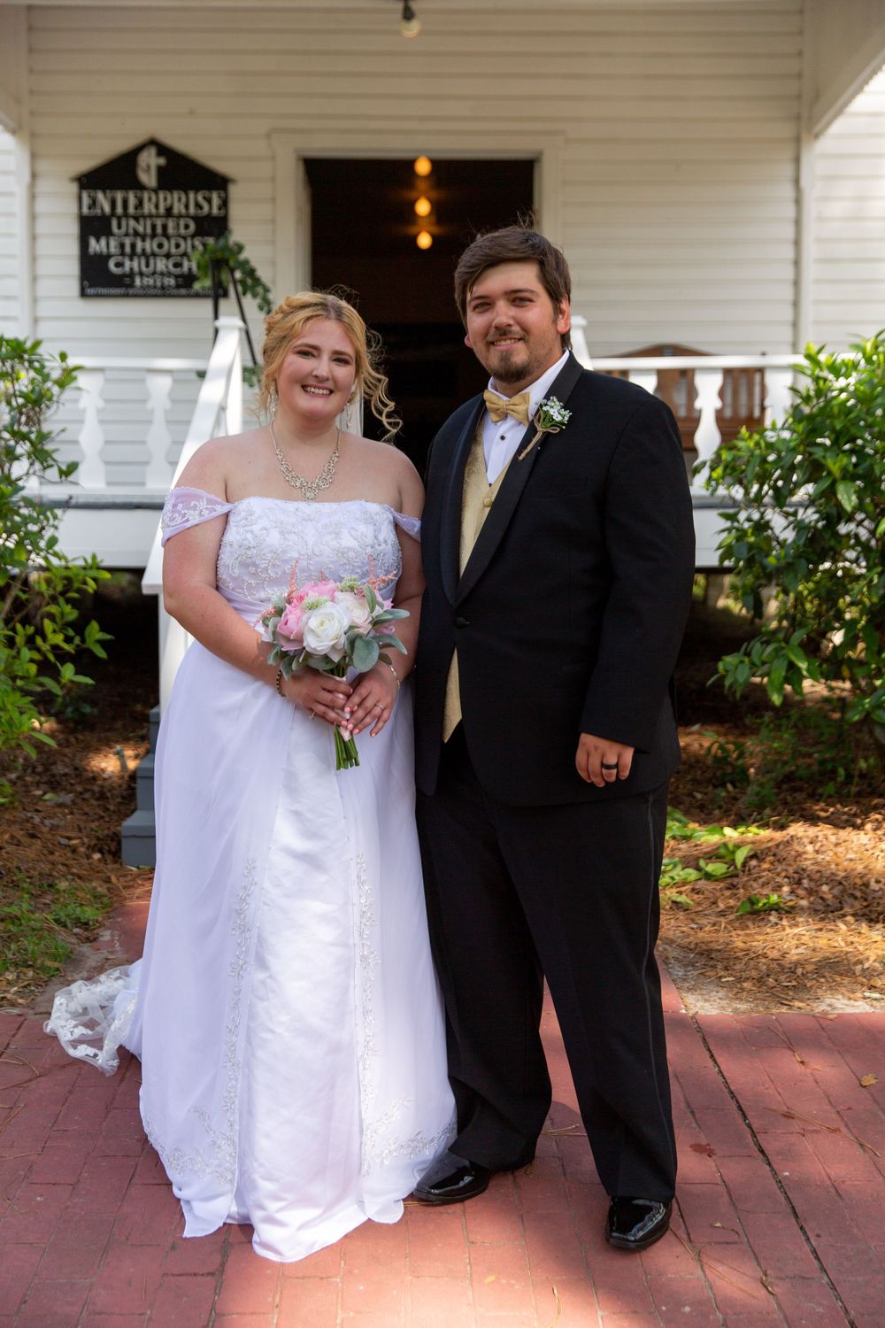 Newlyweds pose together on red brick pathway in front of white church building.