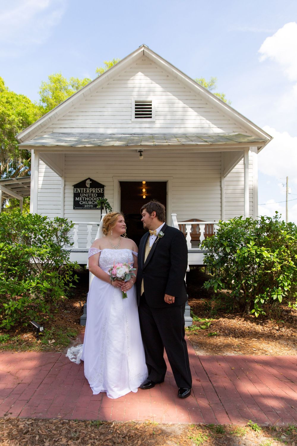 Wedding couple standing together outside white chapel with red brick path.