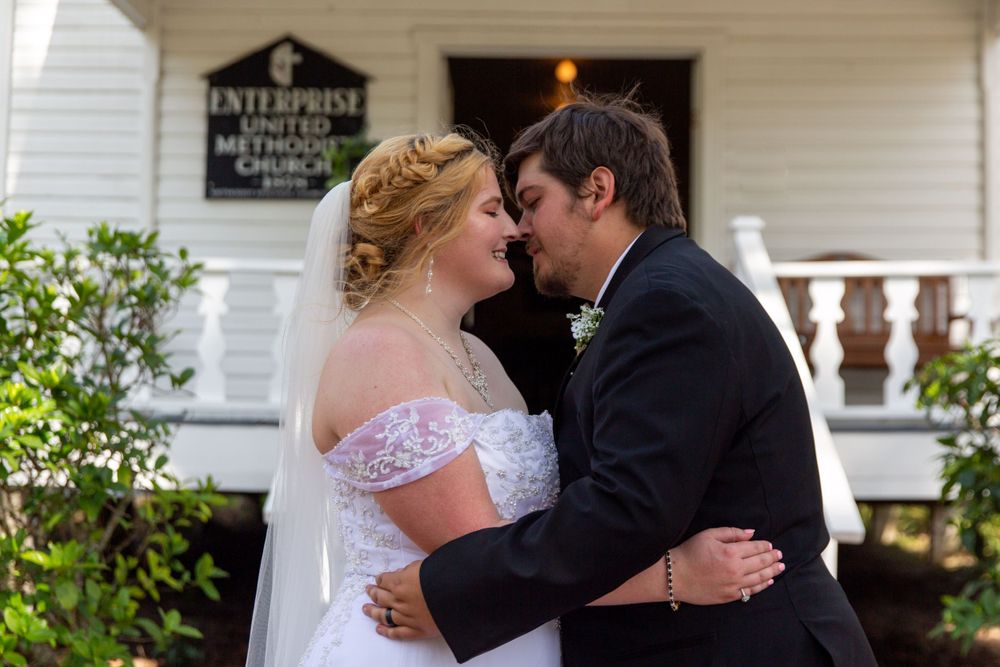 A tender embrace between newlyweds in front of a white building with a pink bouquet.