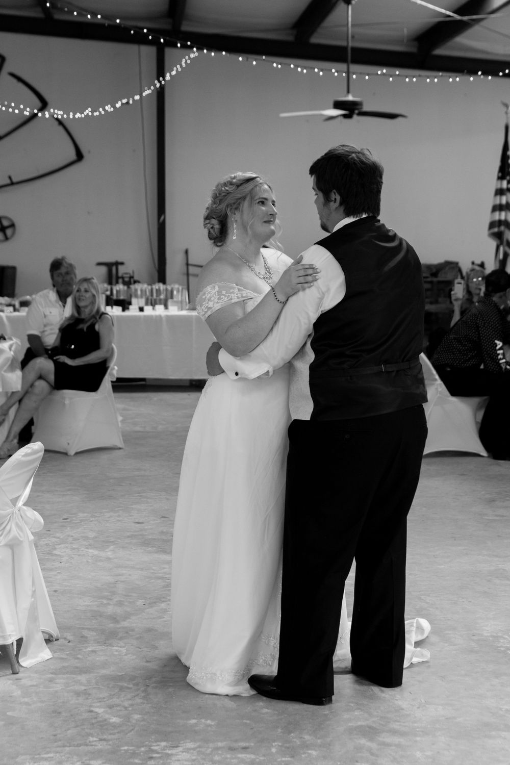 A black and white photo of a couple's first dance at their wedding reception with string lights overhead.