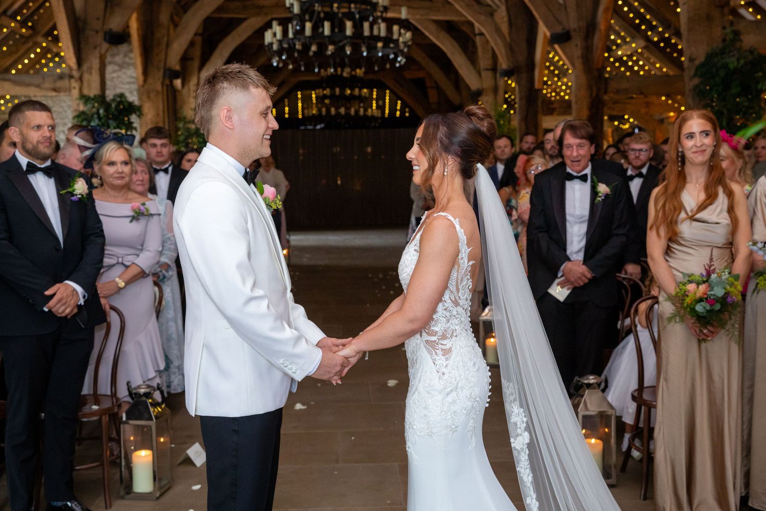 Bride and groom hold hands at rustic barn wedding ceremony with candles and guests.