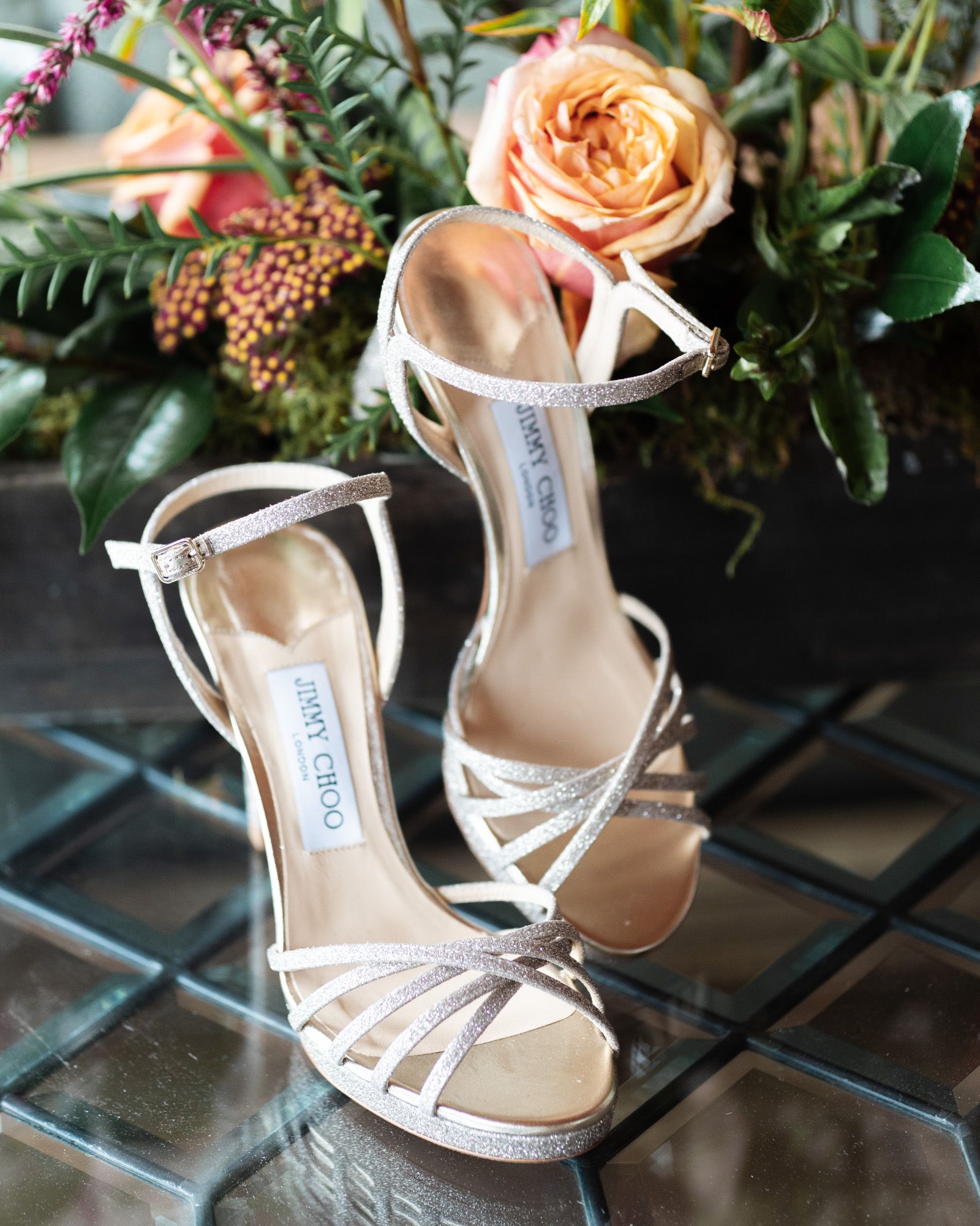 White Jimmy Choo bridal sandals with strappy design displayed with peach roses and greenery on reflective surface.