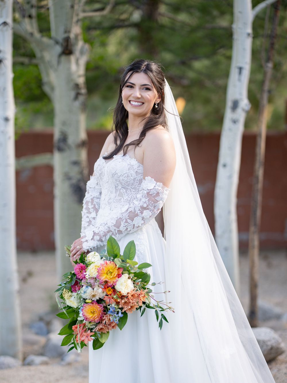 A bride in a white lace dress holds a vibrant bouquet of pink, orange and white flowers against a birch tree backdrop.