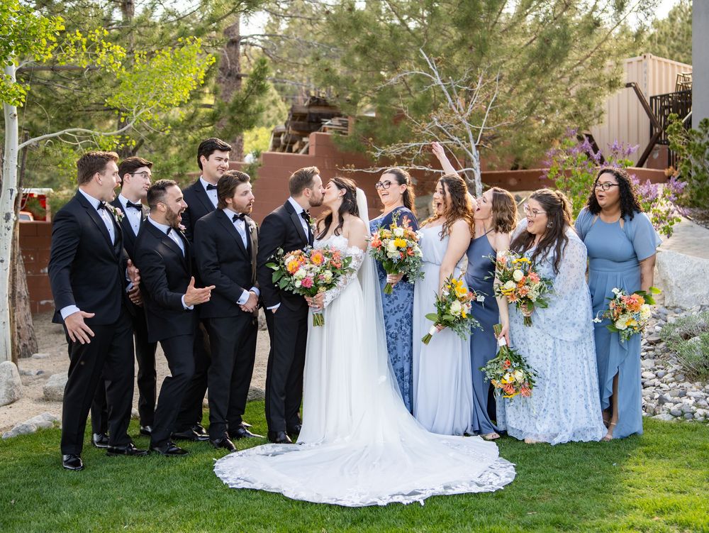 A wedding party in coordinating blue bridesmaid dresses and black suits poses together on a grassy lawn.