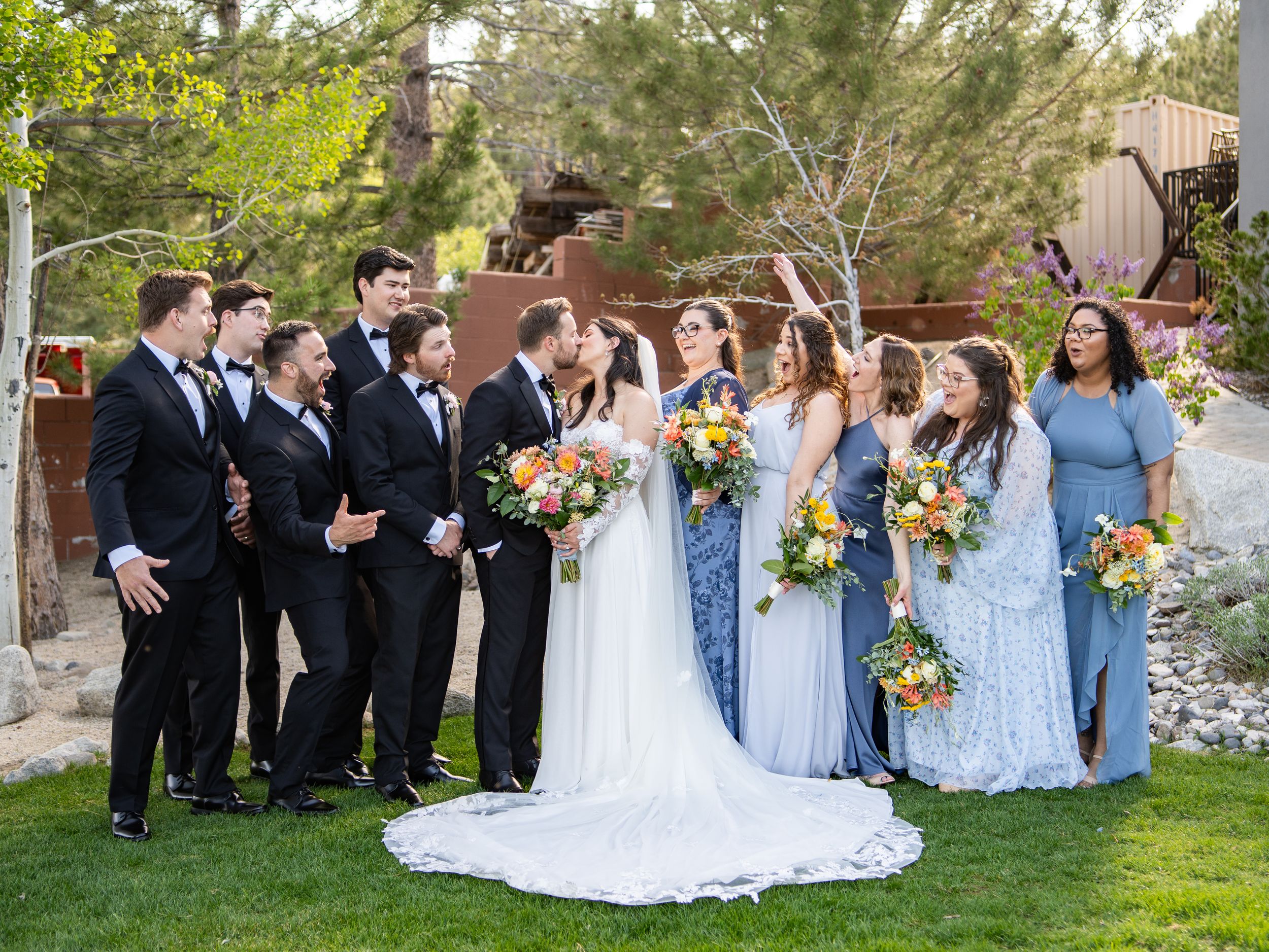 A wedding party in coordinating blue bridesmaid dresses and black suits poses together on a grassy lawn.