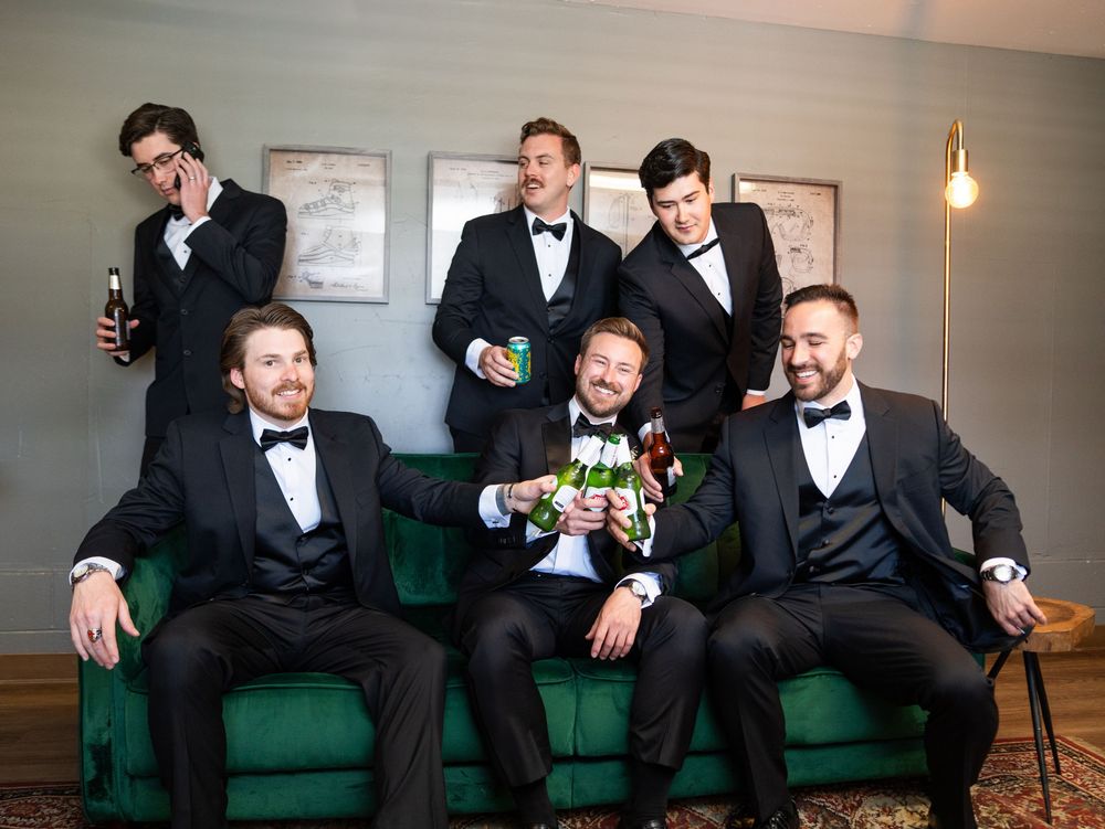 A group of groomsmen in black tuxedos sit casually on green velvet furniture holding beverages.