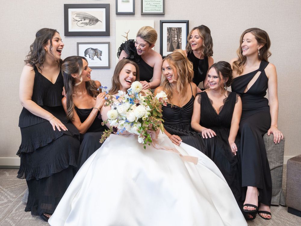 A bride in a white ball gown sits surrounded by bridesmaids in black dresses, all laughing together.