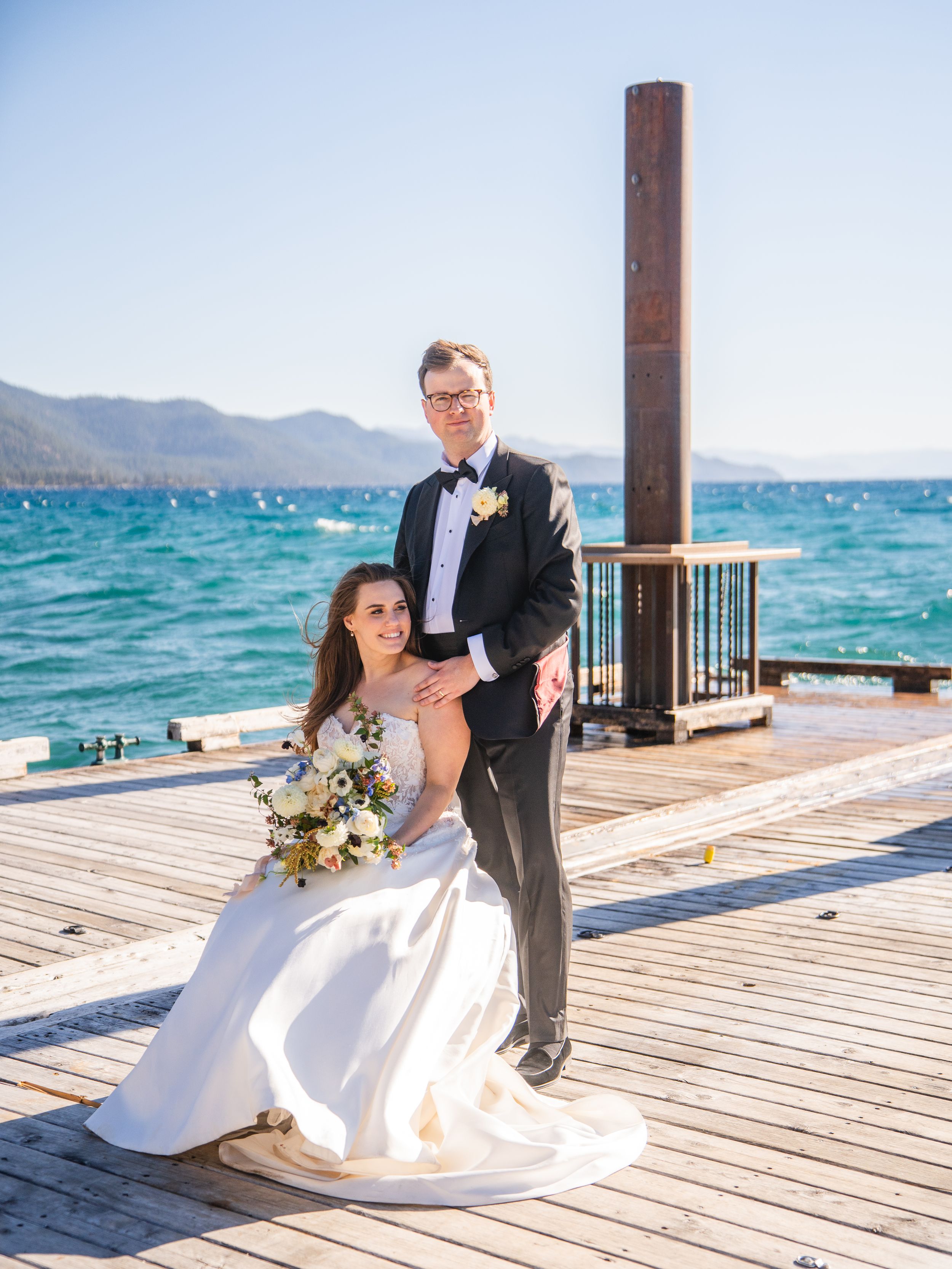 A couple poses on a lakeside dock at Lake Tahoe with mountains and turquoise water in the background on their wedding day.