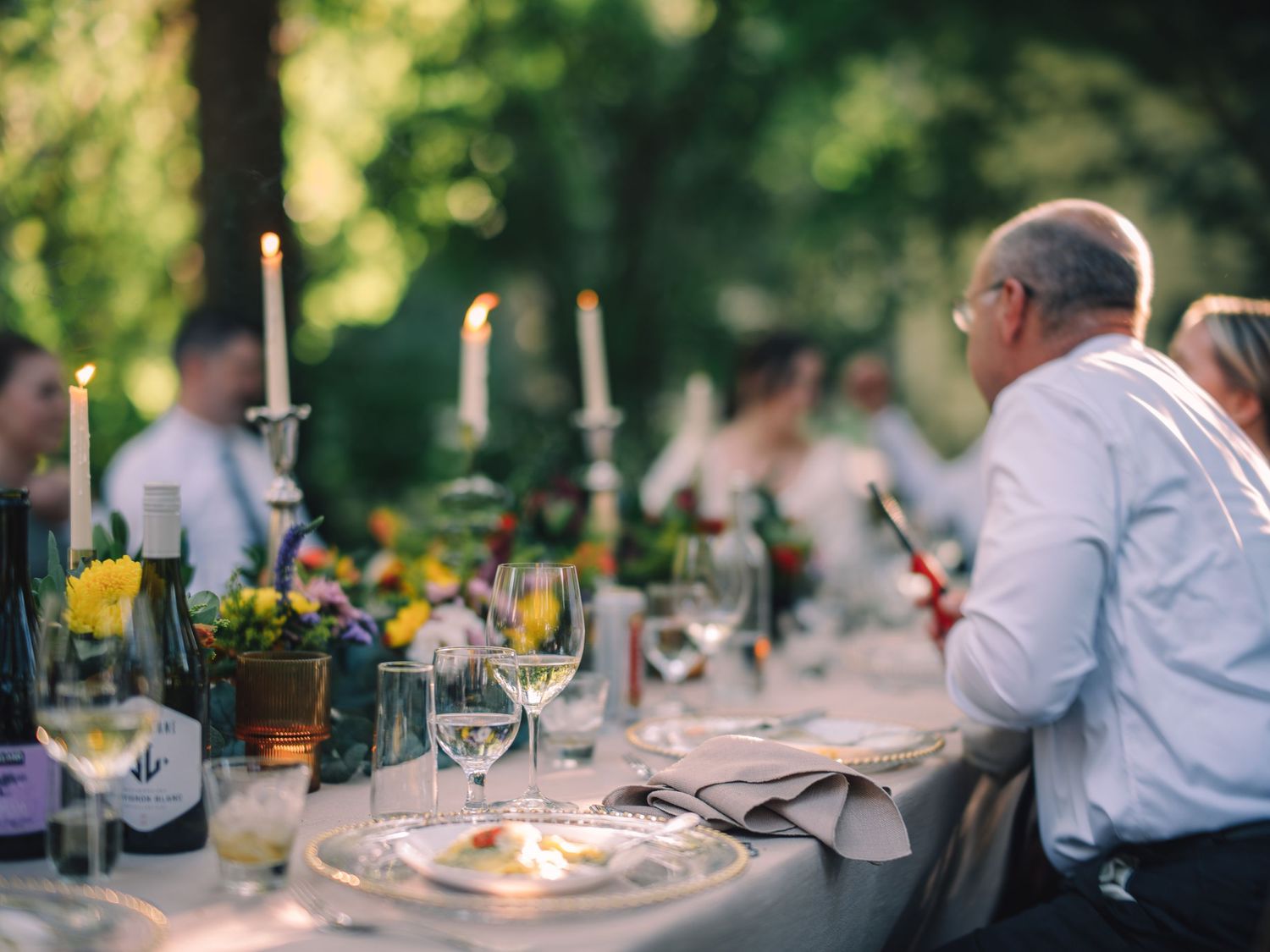 Elegant outdoor dinner party with candles and table settings in a garden setting.