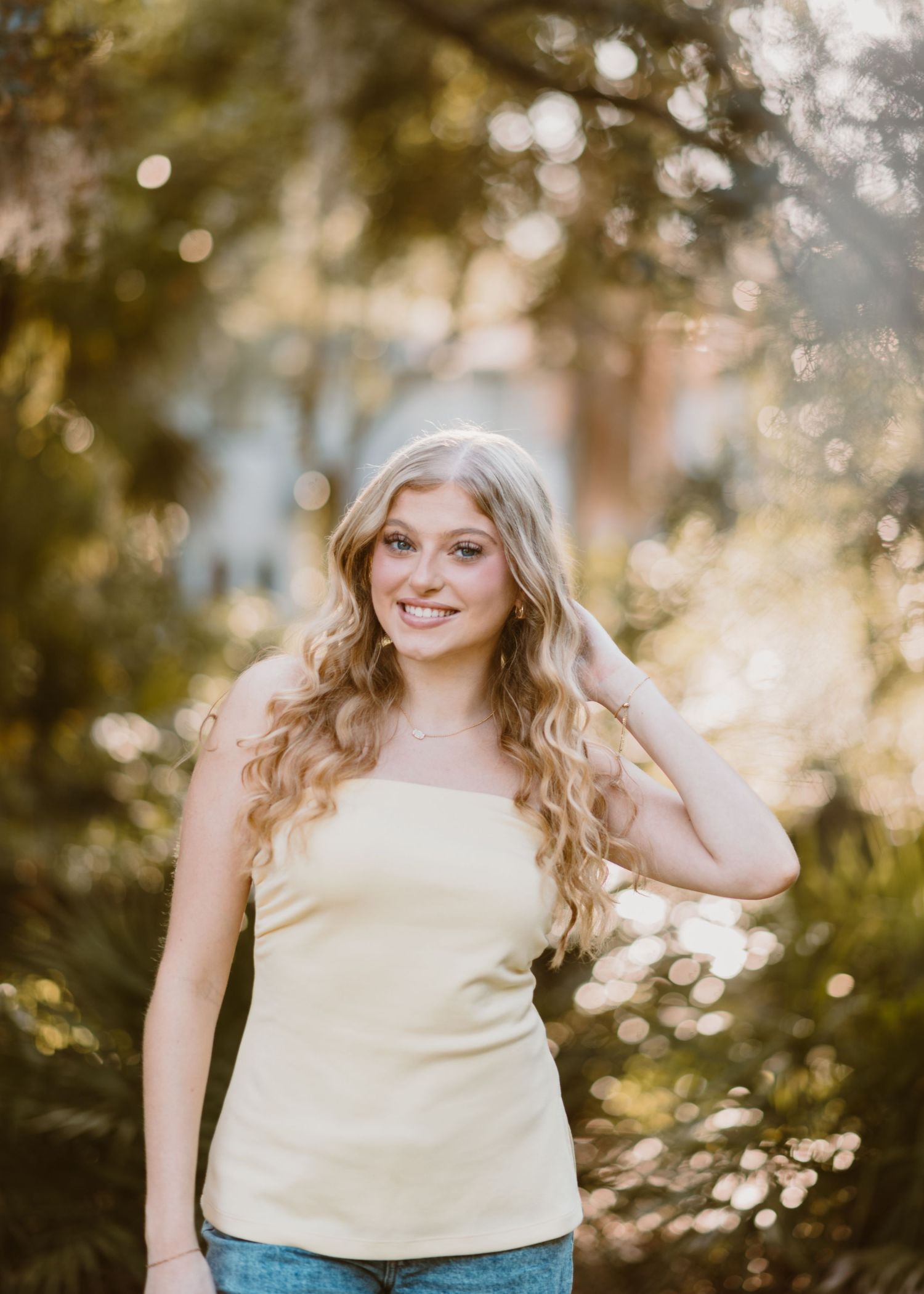 Young blonde girl smiling in a yellow top, with sunshine coming through the trees. Orlando Grad Photos, Dickson Azalea Park Cred: LexTakePix