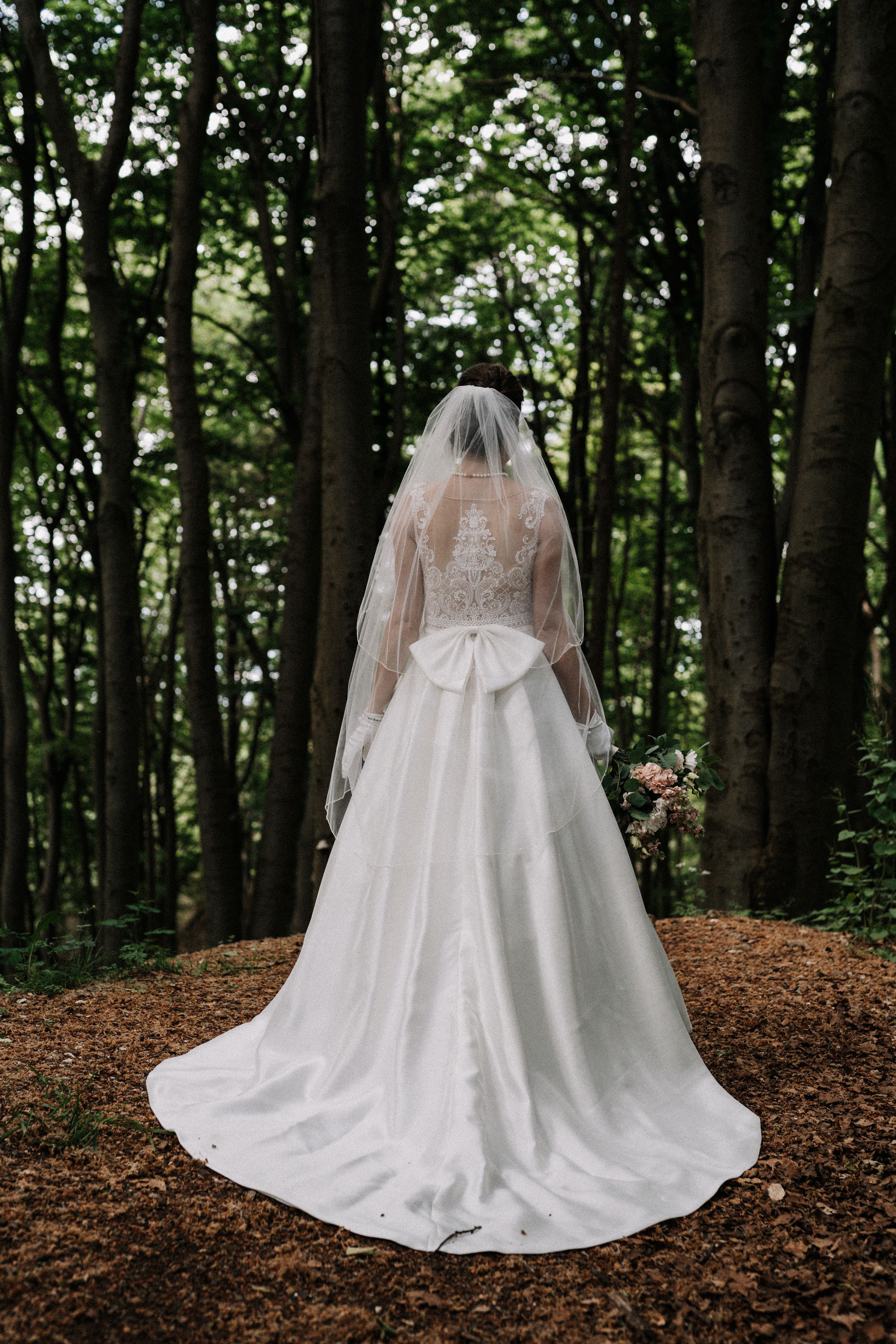 Back view of bride in elegant lace wedding gown with flowing train standing in forest setting.