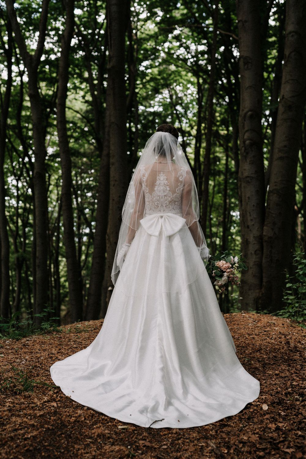 Back view of bride in elegant lace wedding gown with flowing train standing in forest setting.