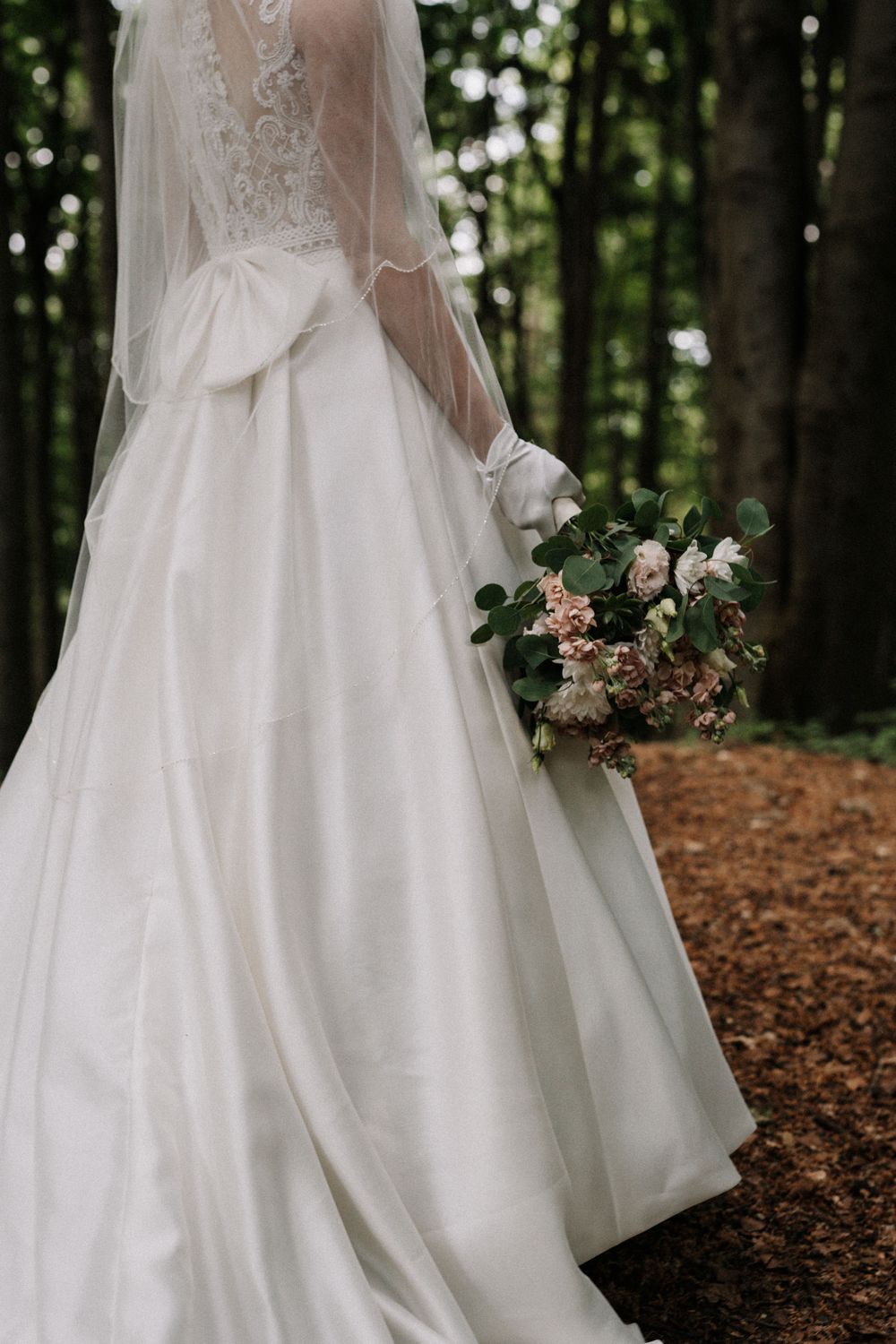 Elegant white wedding dress with lace details and floral bouquet photographed in a forest setting.