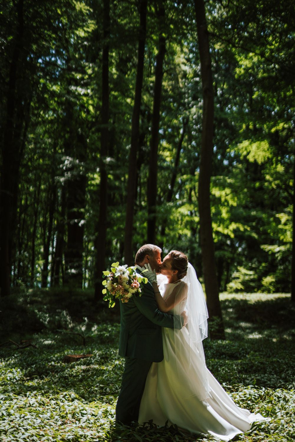 A romantic wedding couple embraces in a sunlit forest surrounded by lush greenery.