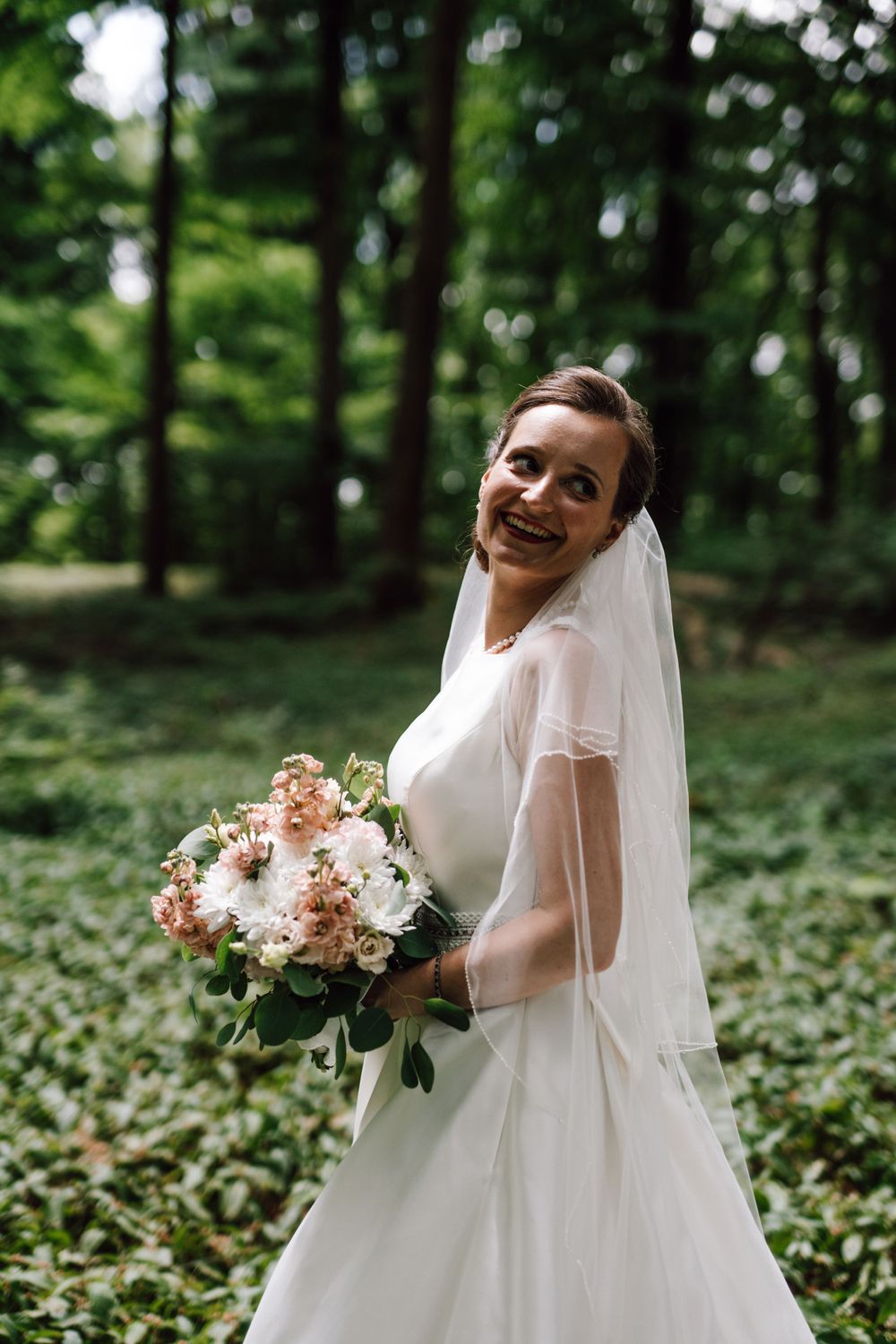 Bride in elegant white dress holds a peach bouquet while laughing in a forest setting.