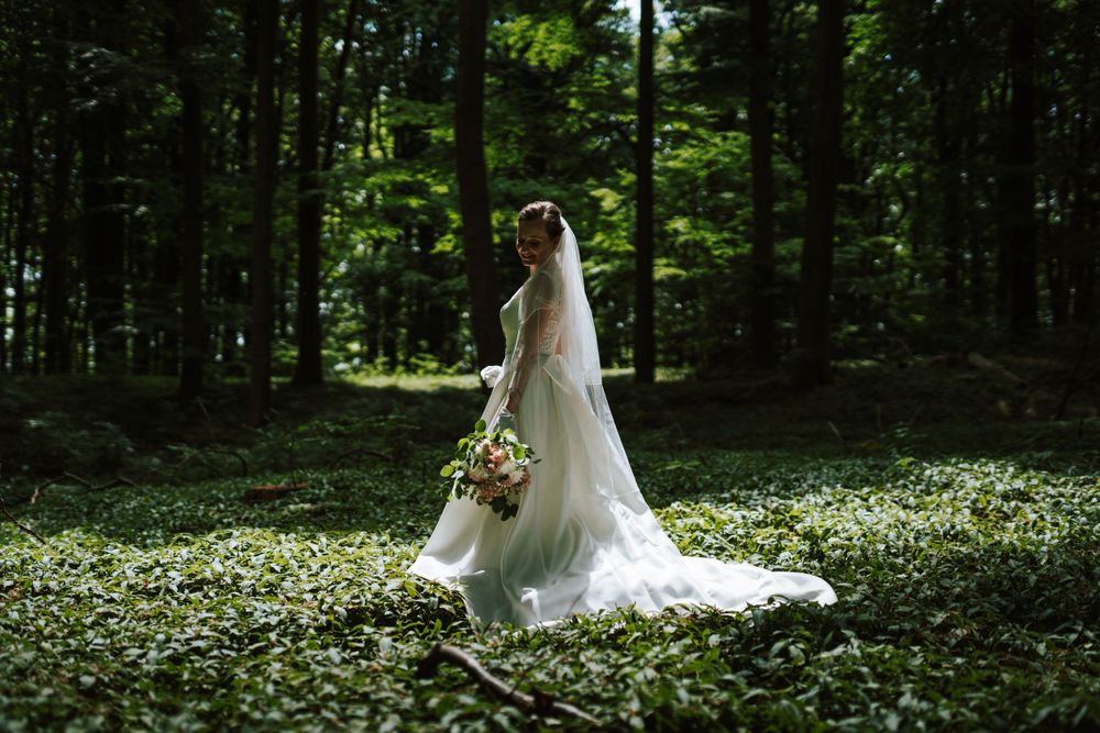 A bride in a flowing white gown stands gracefully in a lush forest setting filled with greenery.