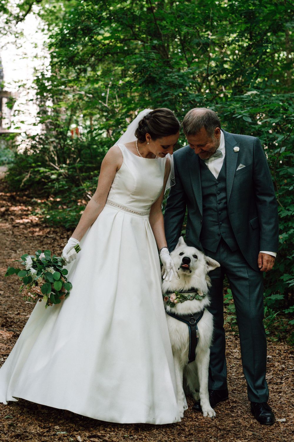 A joyful wedding moment with a fluffy white dog joining the couple in the forest.