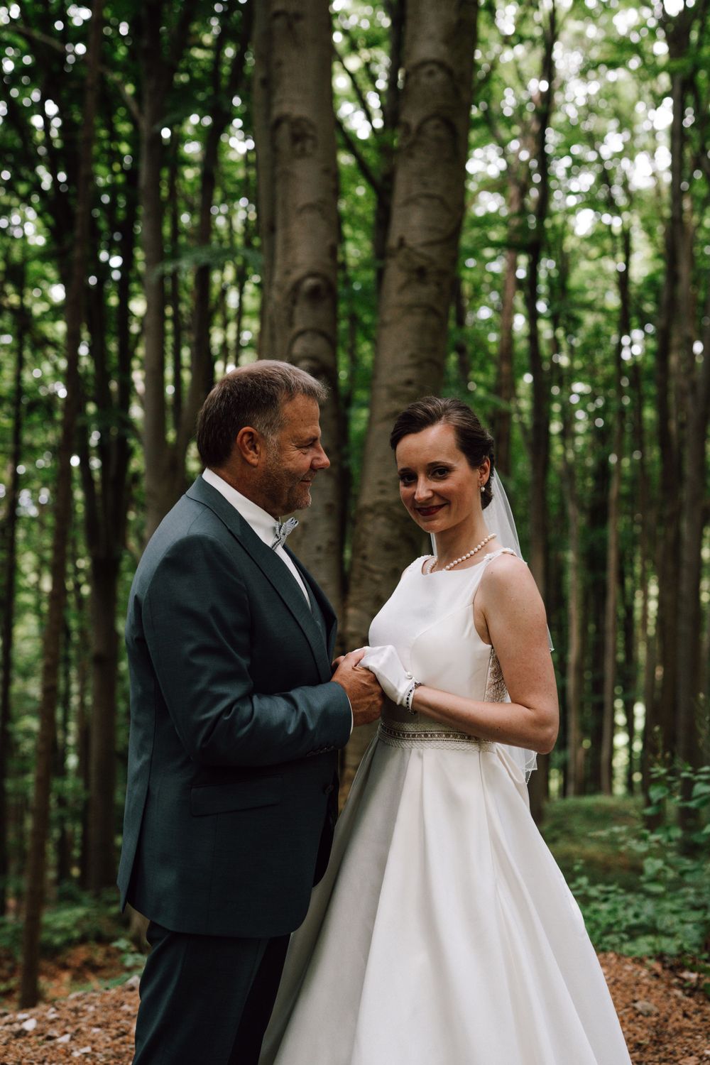 A wedding couple shares an intimate moment in a serene forest setting with tall trees in the background.