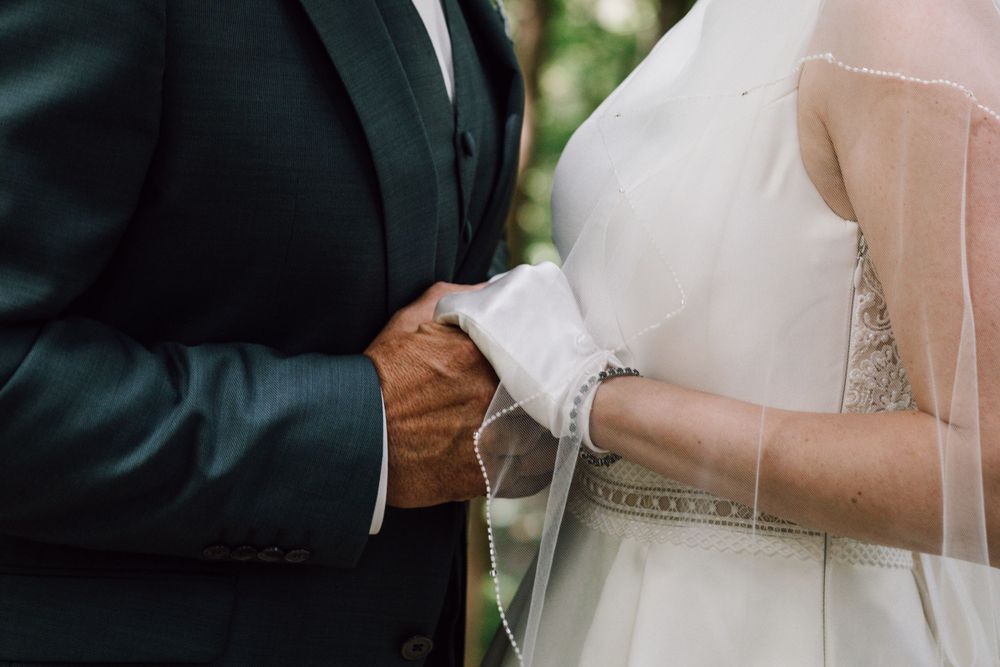 Close-up of newlyweds holding hands during wedding ceremony, with bride in white gloves and groom in dark suit.