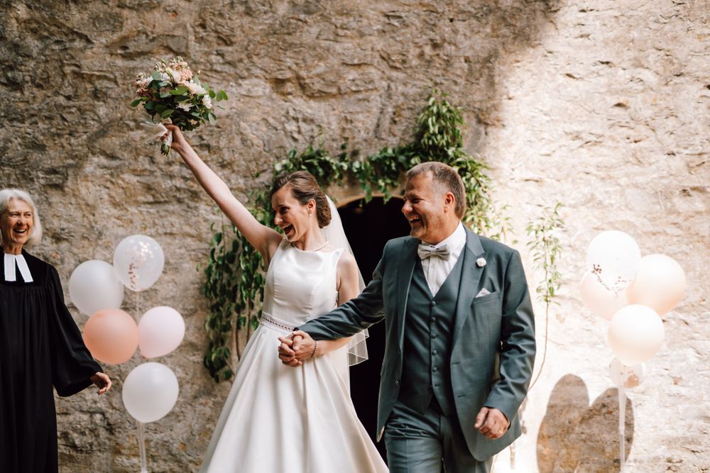 Newlyweds celebrate with raised arms amidst white balloons and greenery decorations.