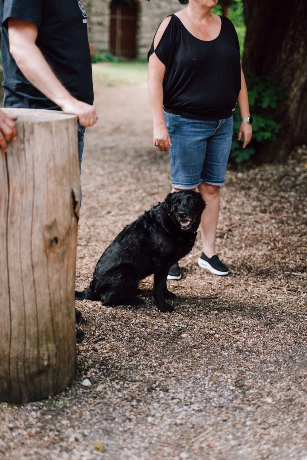 A black dog sits obediently on a gravel path next to people wearing casual clothing.