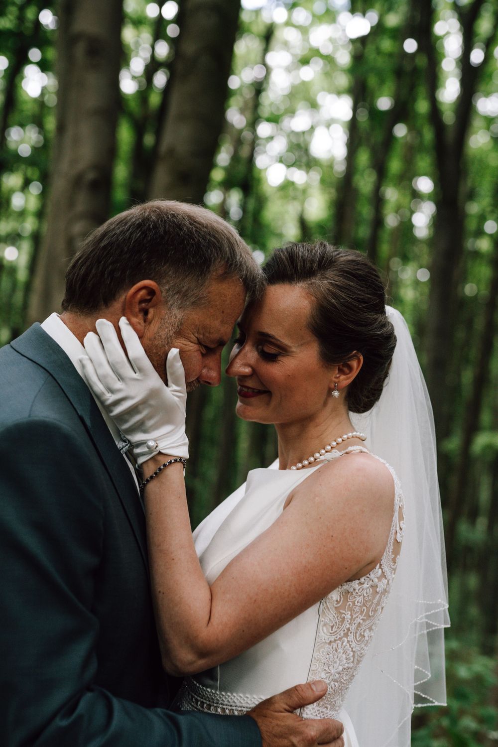 Tender moment between newlyweds in forest setting with bride wearing white gloves and pearl necklace.