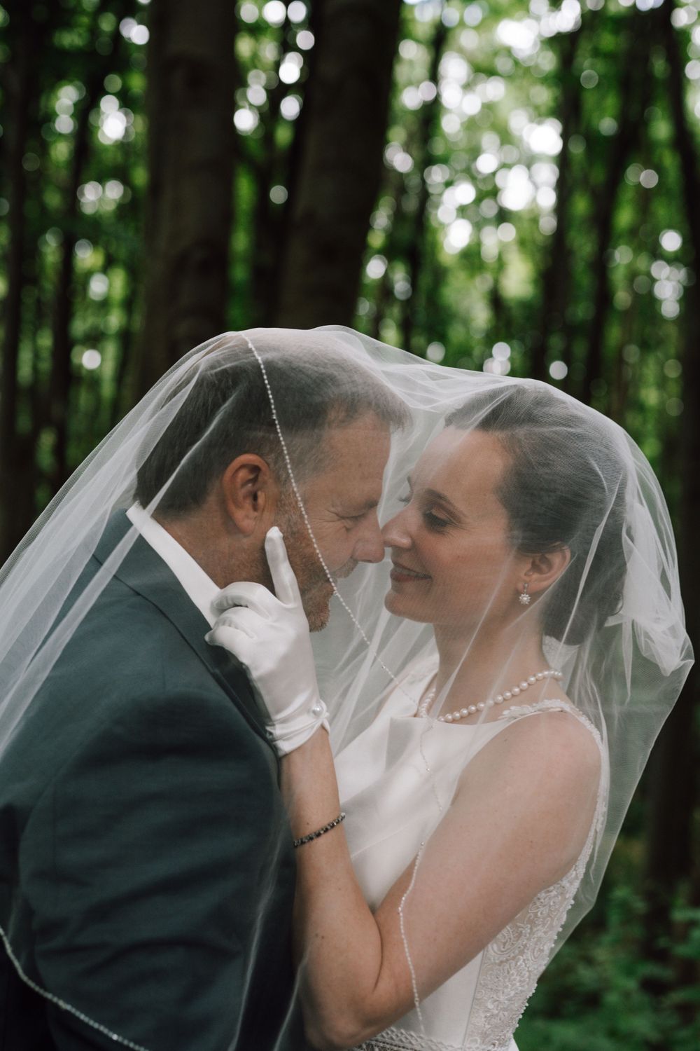 Romantic wedding portrait with couple sharing intimate moment under flowing veil in forest setting.