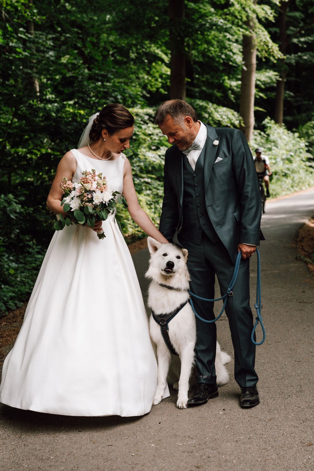 A white fluffy dog poses with a couple on a tree-lined path.