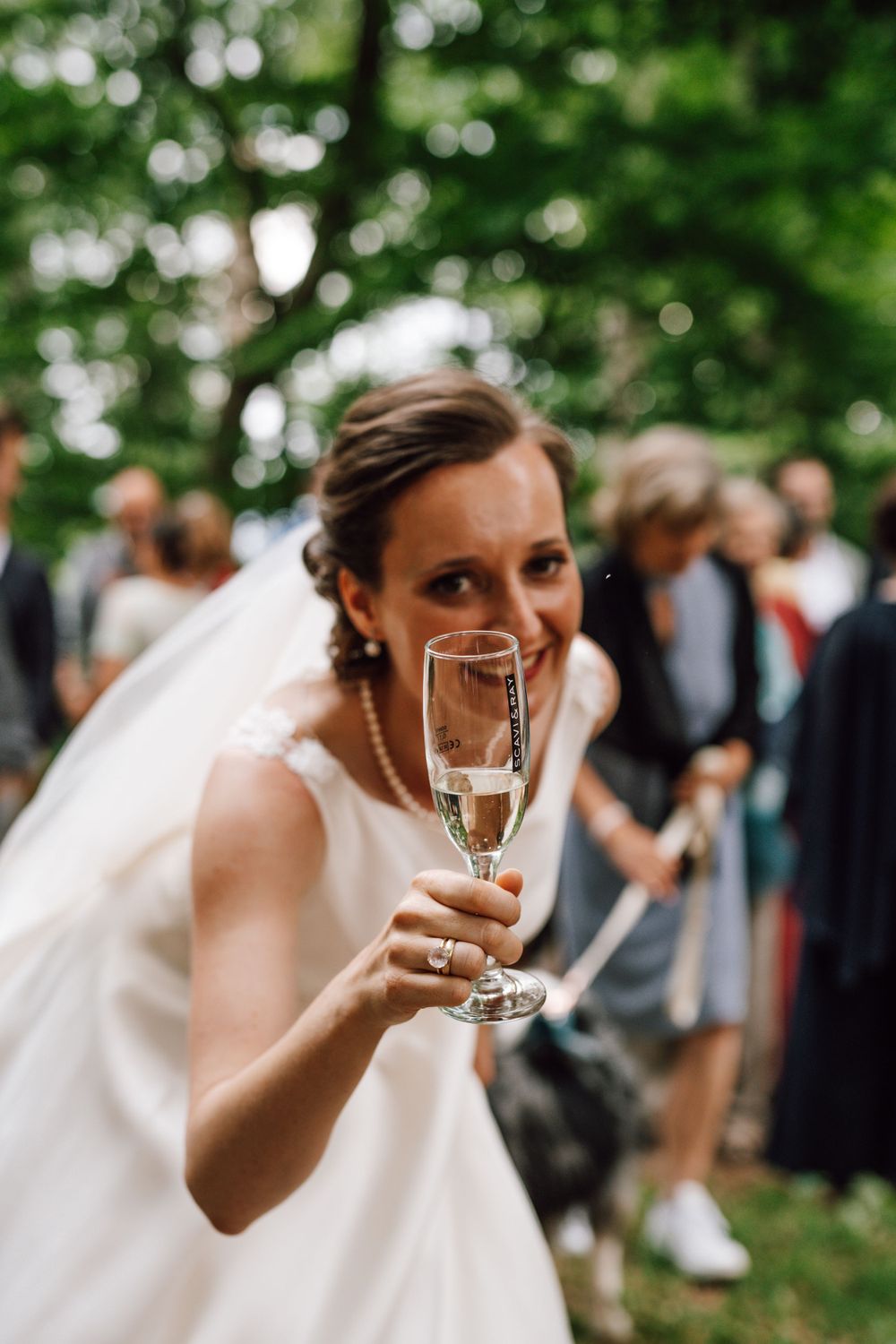 A person in a white wedding dress holds up a champagne glass while smiling outdoors.