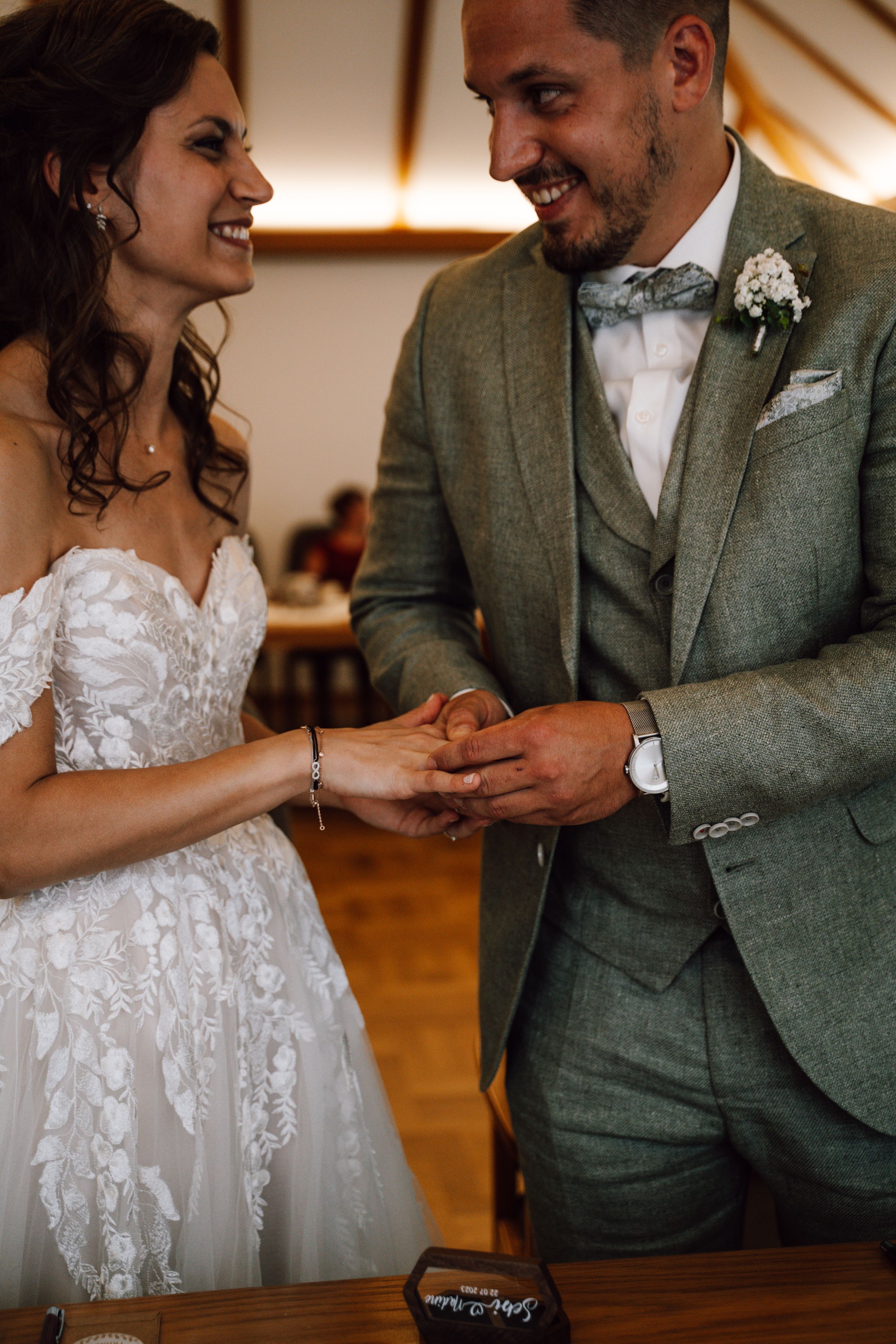 Intimate moment of ring exchange between couple in lace gown and sage green suit.