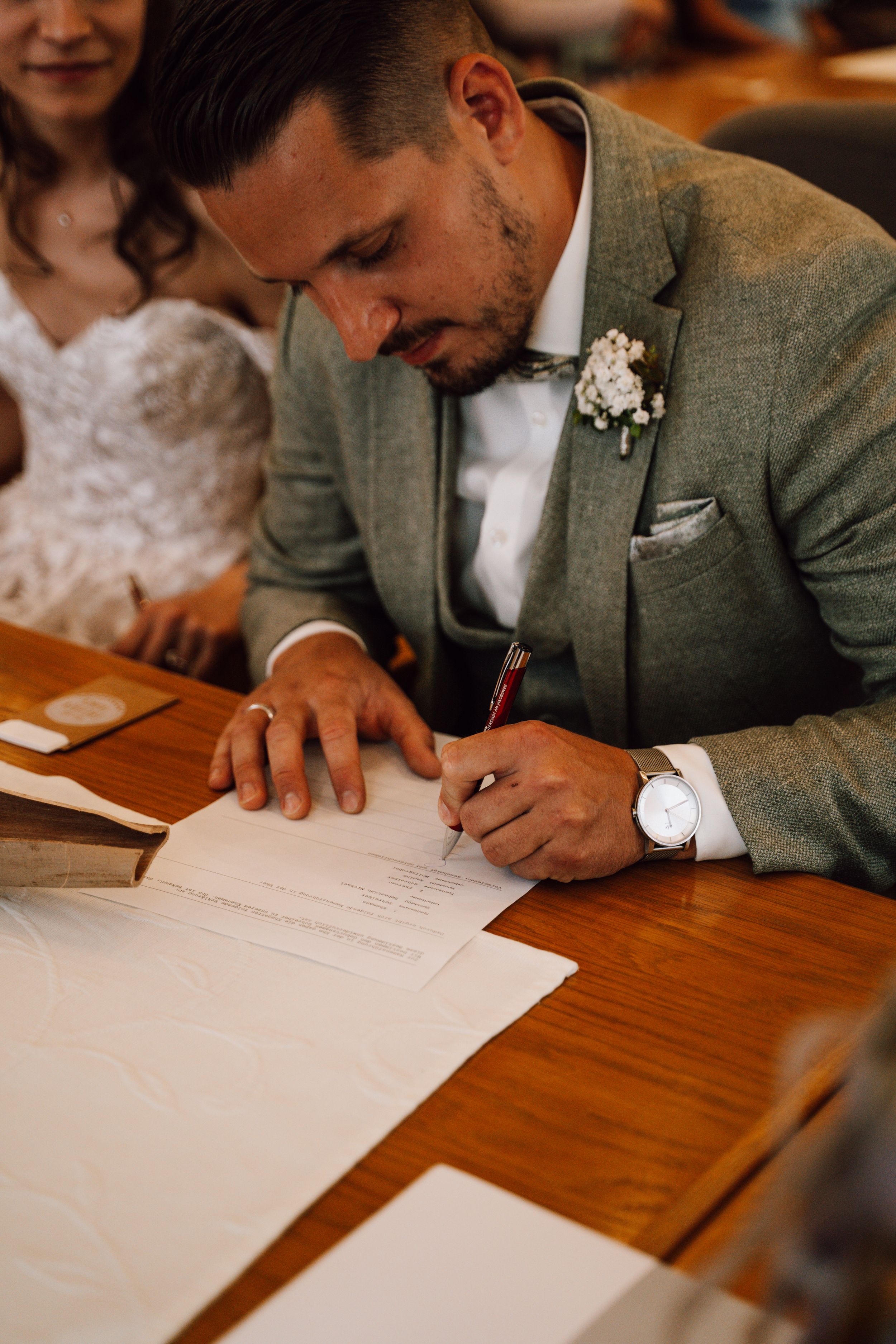Close-up of someone in a gray suit signing documents on a wooden table.