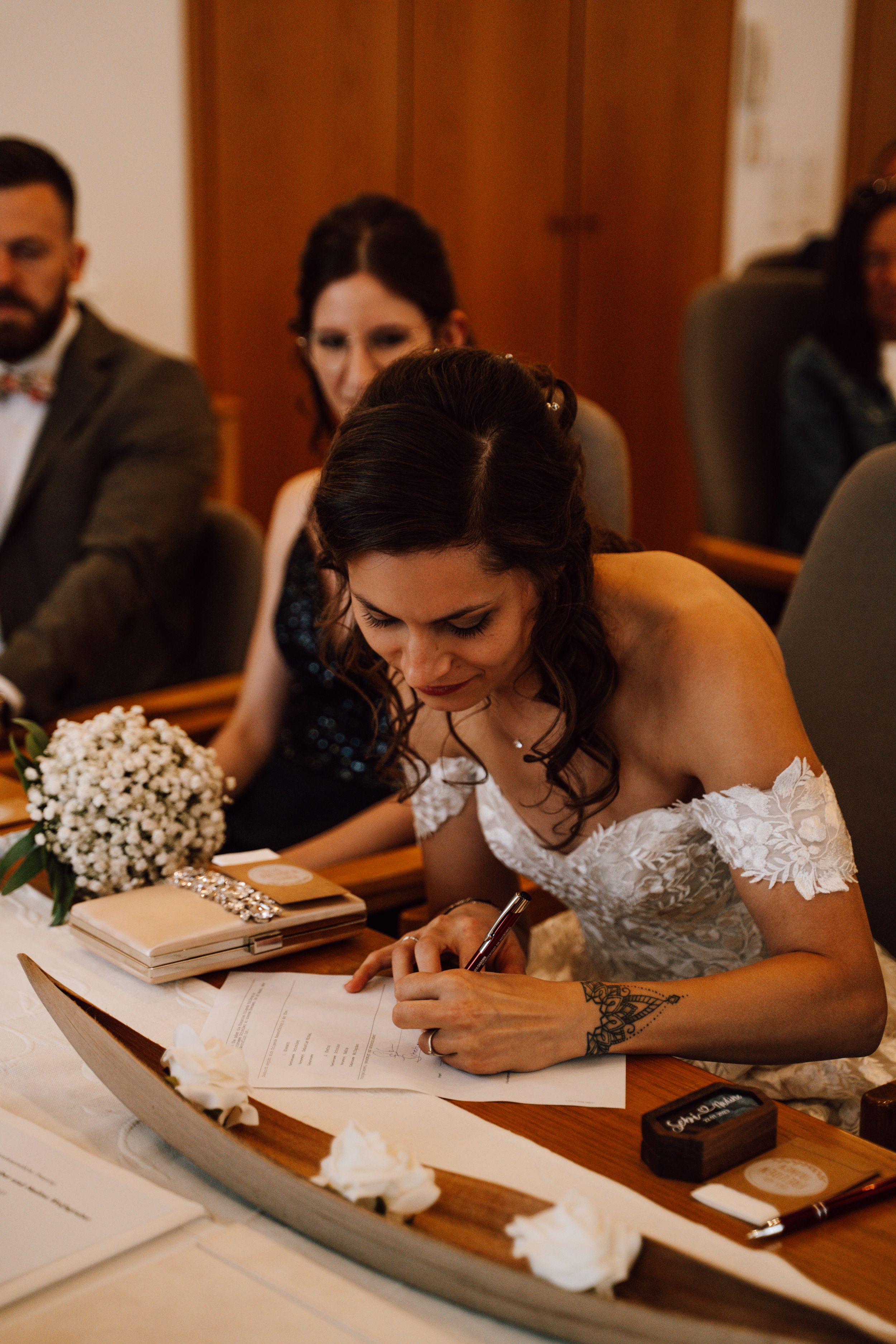 Bride in a white lace dress signing wedding documents at a wooden table.