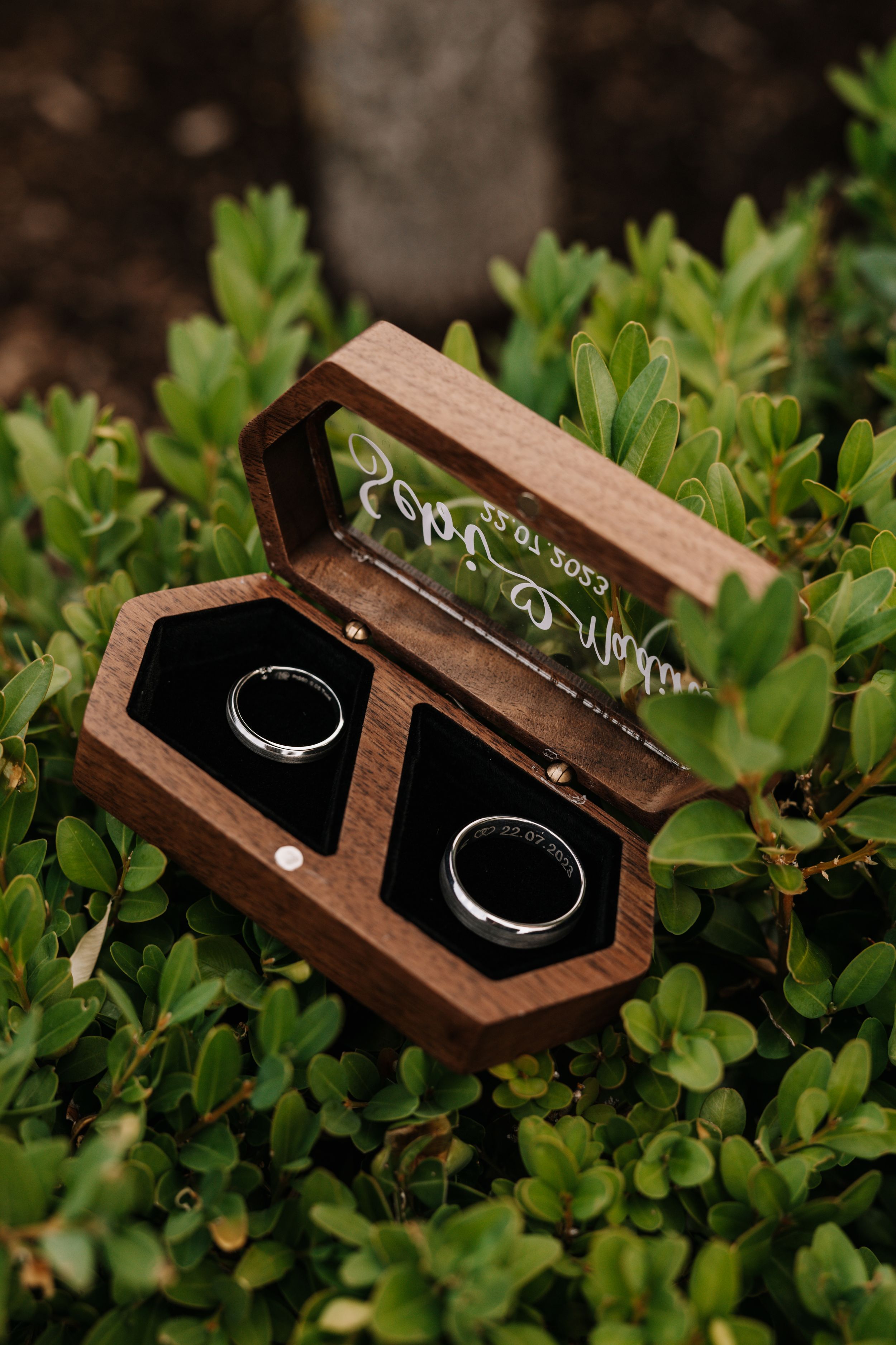 Wedding rings displayed in a rustic wooden box nestled among green foliage.